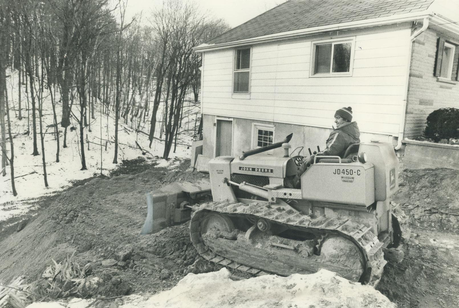 A small bulldozer moves earth from the basement of a small house, next to a woodlot.