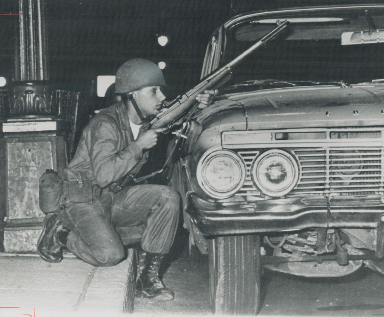 Sniping at Snipers, a national guardsman crouches behind a parked car last night outside 10th district police station, on Detroit's west side, trying (...)