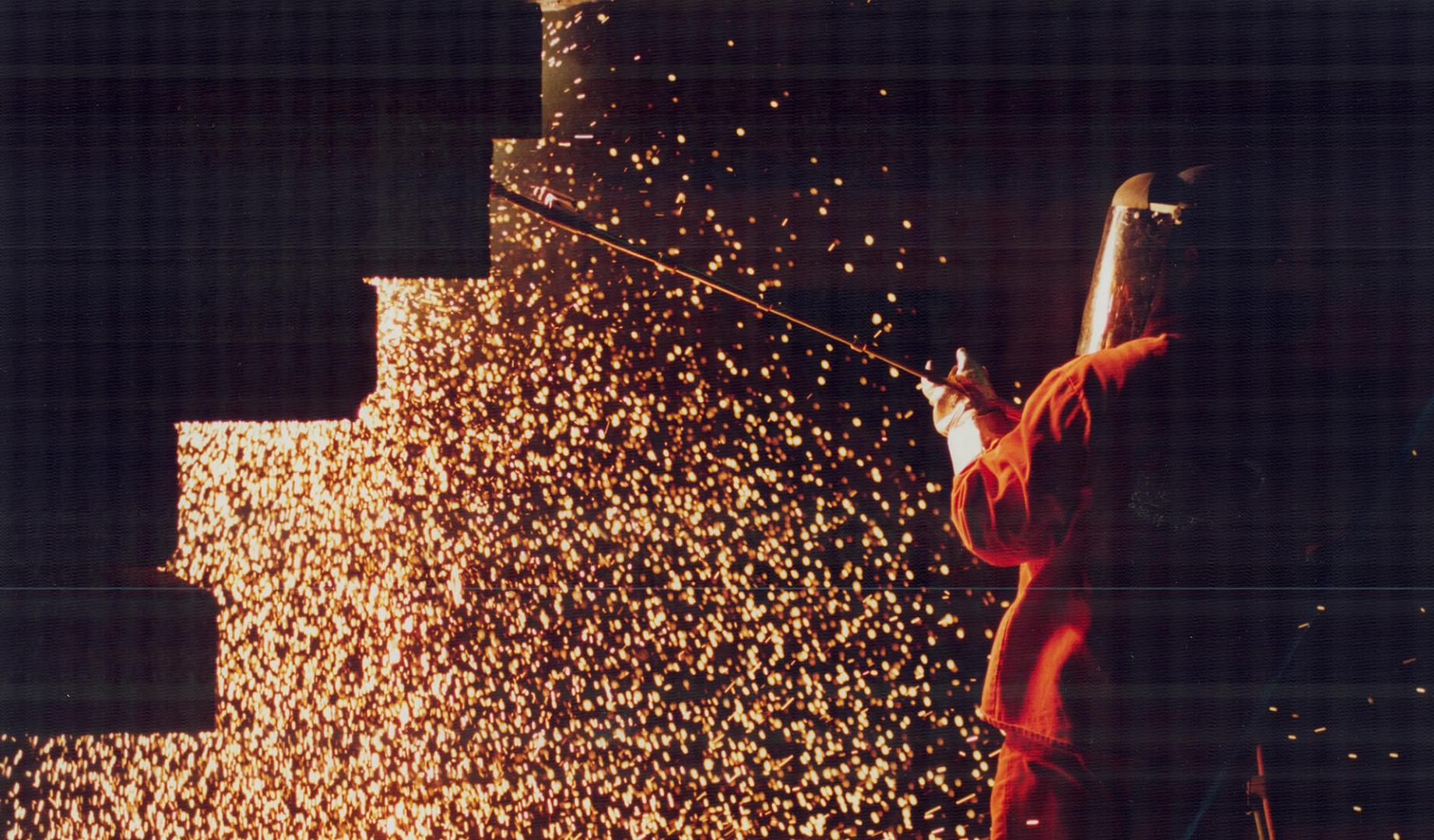 A man in an orange flame-retardant suit and visor uses a tool next to a shower of red sparks.