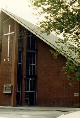 Image shows a part of the church building front side with a church sign and a big cross closer …