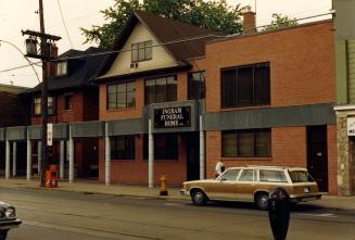 Image shows the street facing side of the funeral home and a car parked in front of it.