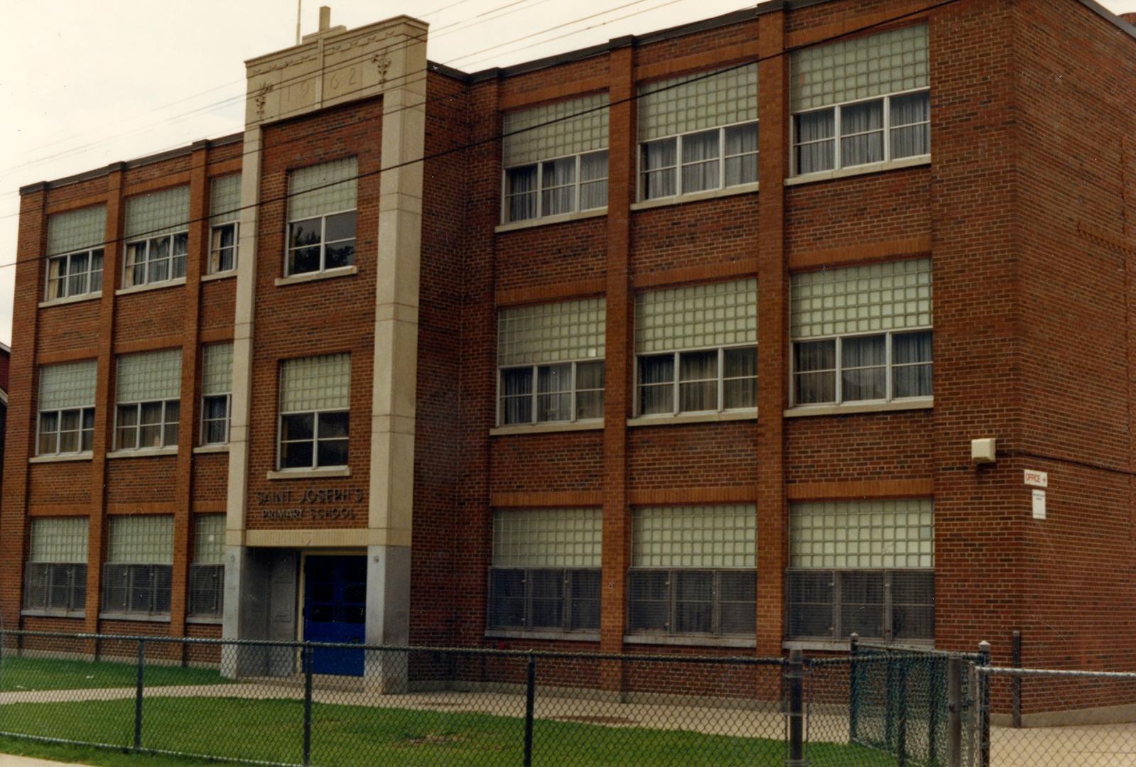 Image shows a three storey school building with a fence around from the main entrance side.