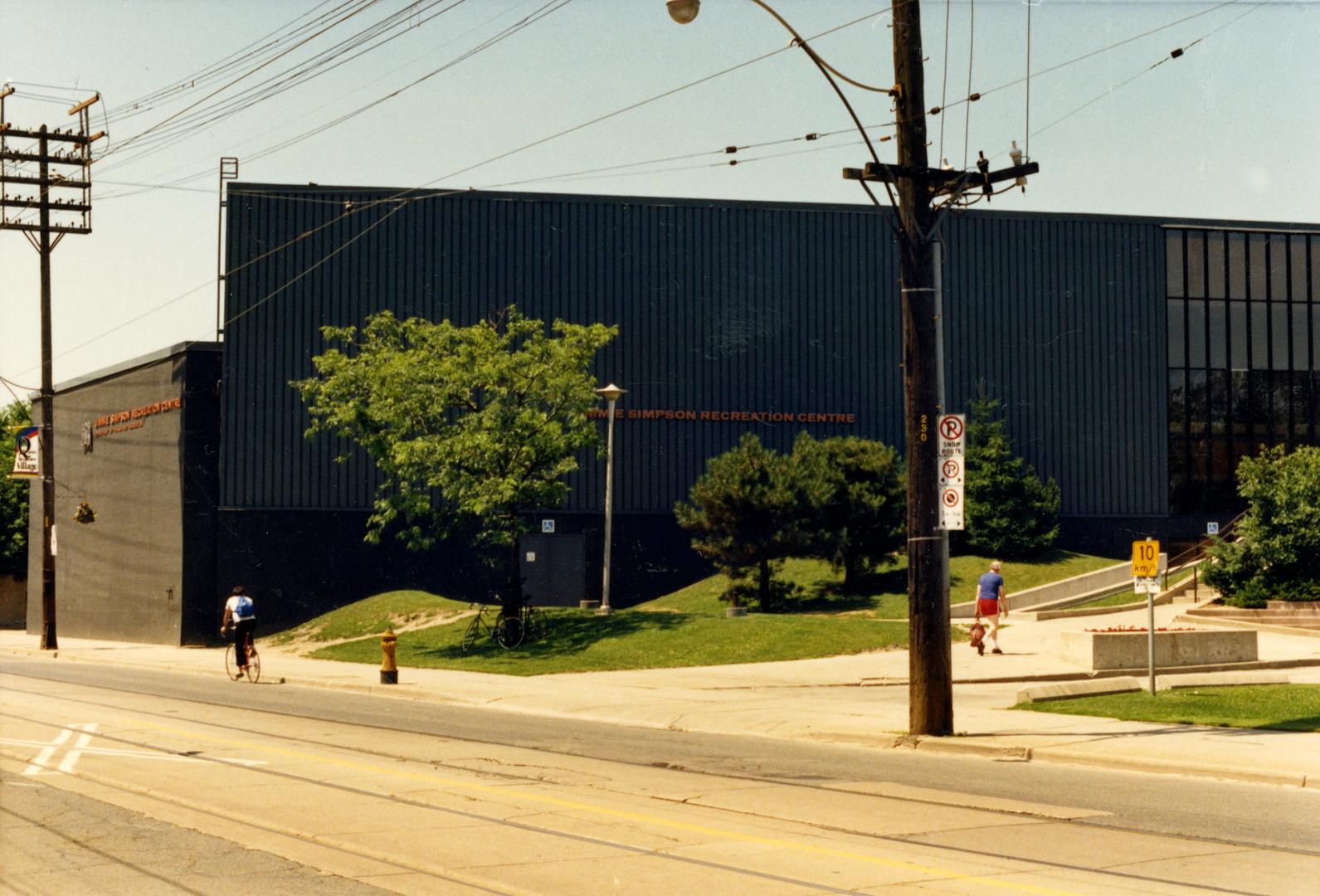 Image shows a street view with a recreation centre on one side. There are some trees and bushes…