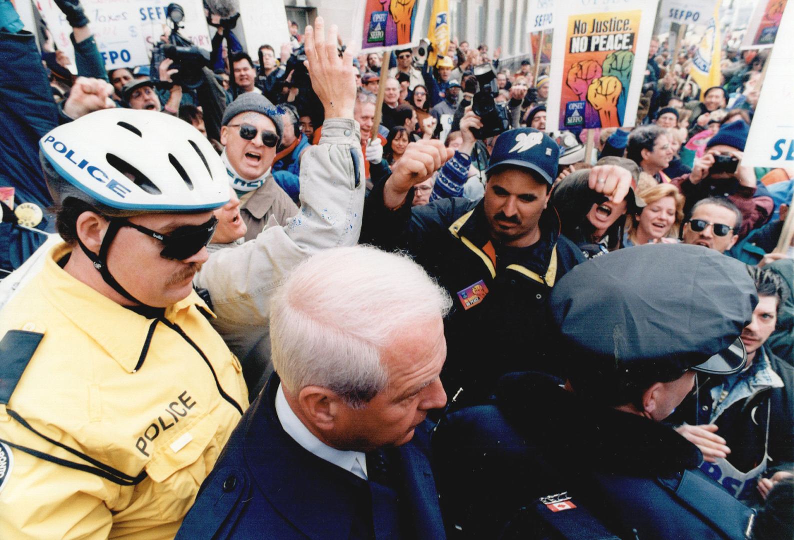 Reporters surround a man in a blue suit, who has two police escorting him. Protesters with sign…