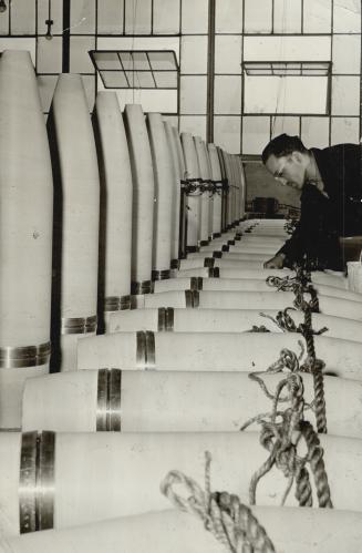 These Big Shells being painted by John Havryluk for rust protection, are a small drop in the vast flood of Made-in-Canada munitions pouring, says Hon. C. D. Howe, into all war theatres