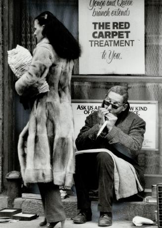 Underneath a bank window sign on Queen St. East near Yonge St., a mouth-organist plays for donations from passersby