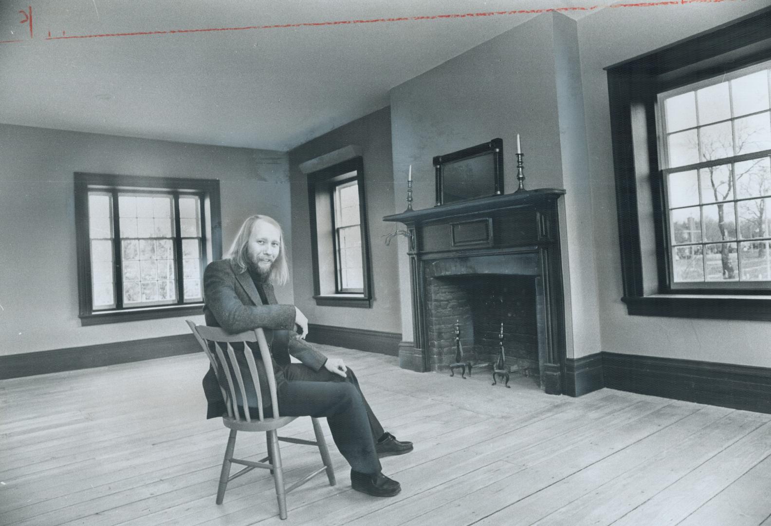 A man sits on a wooden spoke-back chair in centre of empty room with bare hardwood floors and d…