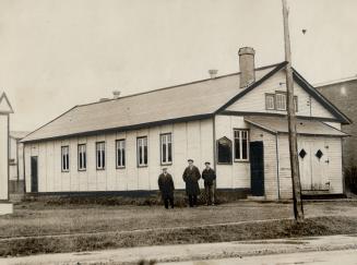 Three men in caps and long dark coats stand in front of a two-storey white clapboard structure  ...