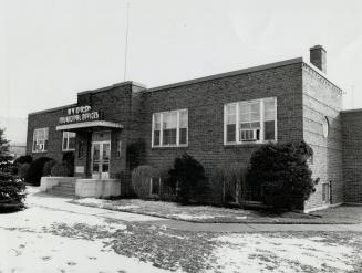 Exterior view of one-storey, flat-roofed brick building with shrubs planted either side of entr ...