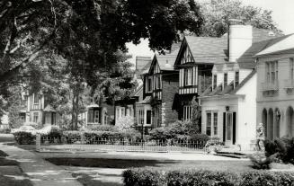 View from the sidewalk of row of homes with large lawns and tidy shrubs in front. The canopy of…