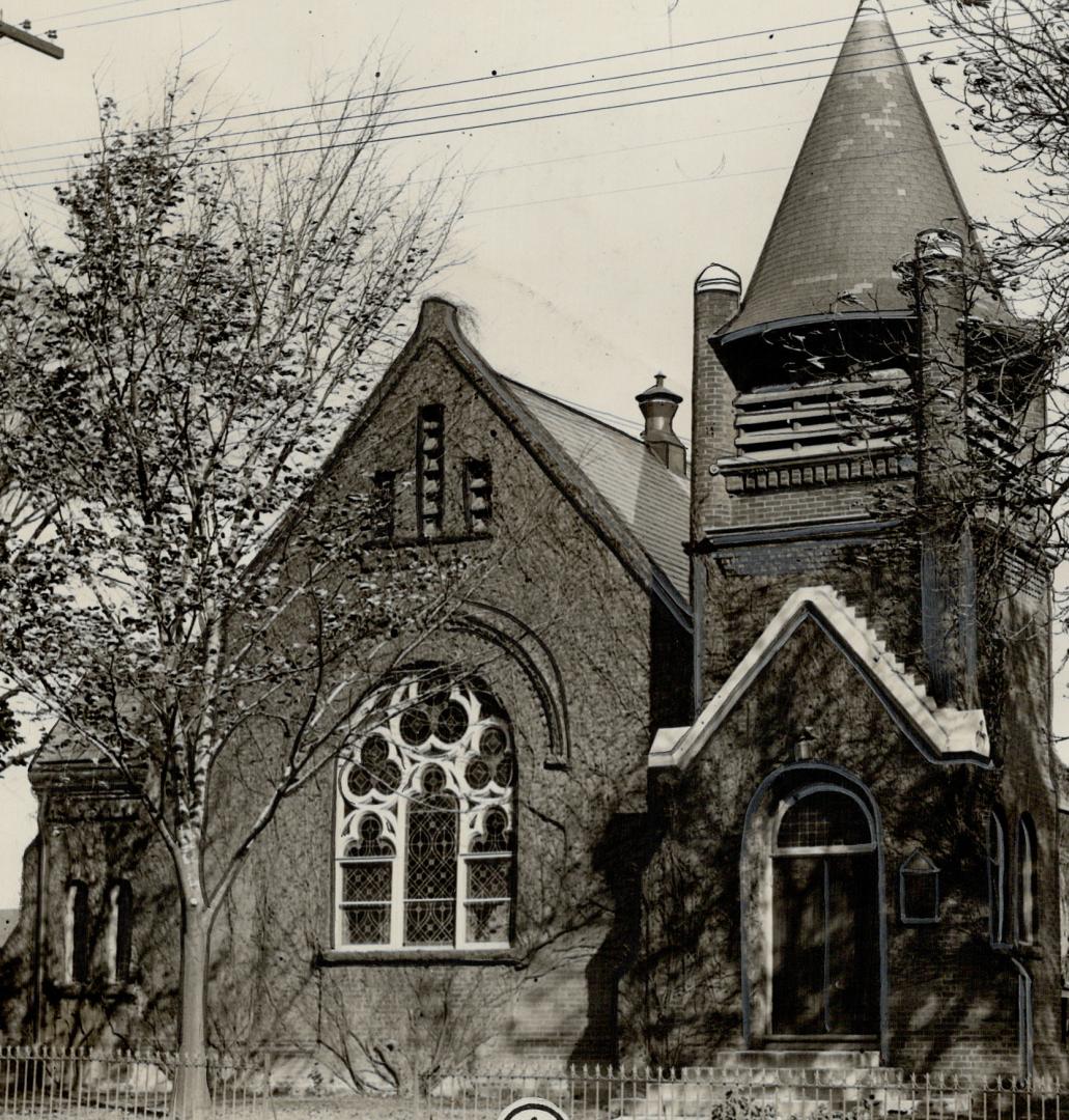 Brick church with steeply pitched gable roof, left, and trefoil window. Belfry tower with turre…