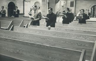 Men dressed in suits (one in religious habit) stand sparsely occupied pews with songbooks in ha…