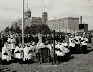 Archbishop wearing robe and decorative ceremonial headgear stands before a black cross on lawn …