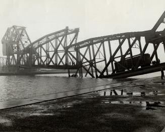 A train trestle is broken in two, collapsed into a river, with a single train car and several p…