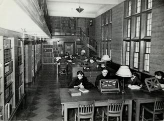 Men seated with books open at wooden tables with lamps and reading stands. Bookshelves line the…