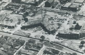 Aerial view of construction site on parcel of land between two roadways, with houses and low-ri…