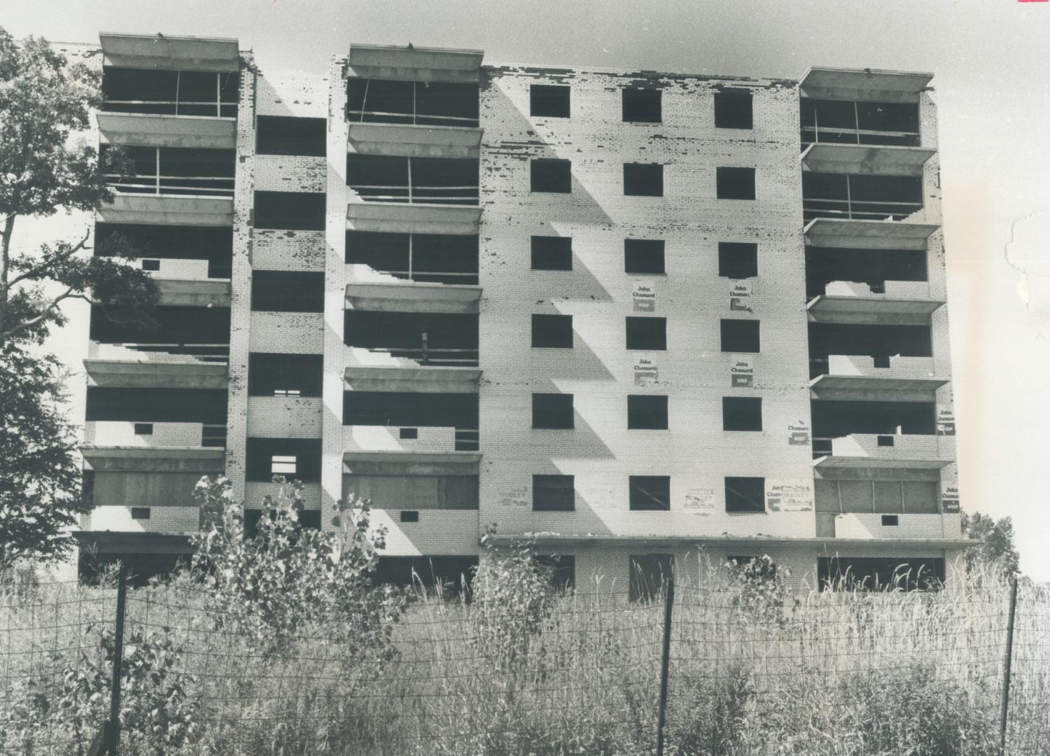 Seven-storey brick building with worn, white paint, missing balcony rails and windows. Affixed …