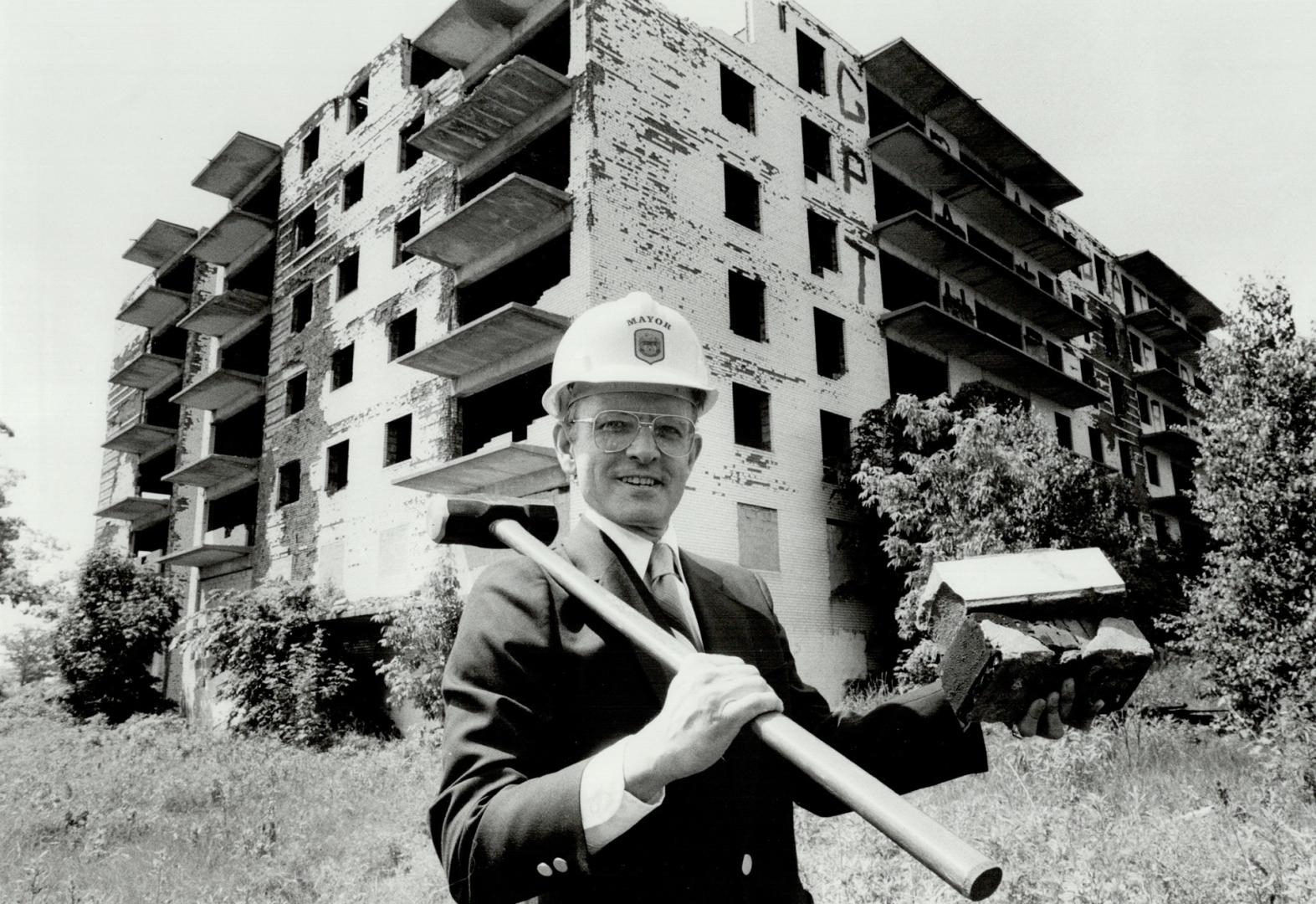 Man wearing suit and hardhat smiles as he rests sledgehammer over shoulder and holds up brick; …