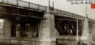 View from below of a large bridge, showing cement support pillars and steel arches. Decorative …