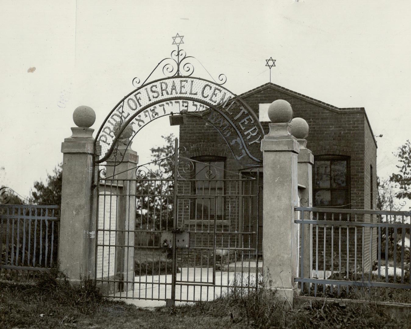 A small, one-storey brick structure stands beyond an arched entrance gate displaying the words  ...