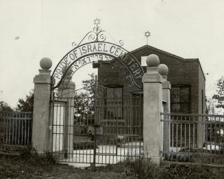 A small, one-storey brick structure stands beyond an arched entrance gate displaying the words …