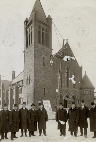 Nine men in long dark coats and fedoras stand in a row facing camera. In background, is a churc…