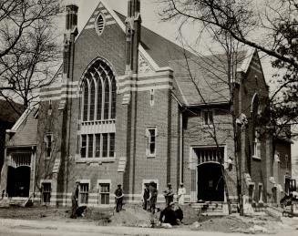 Men with shovels stand and kneel around a mound of earth in front of a church. 