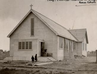 Exterior of unadorned, white clapboard church with small cross affixed to roof and several entr…