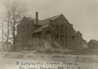 Exterior of brick church on elevated grounds, with cross gable roof, tall narrow chimney stack …