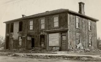 A two-storey structure in disrepair with shingle roof and brick chimney. Sign affixed to wall r…