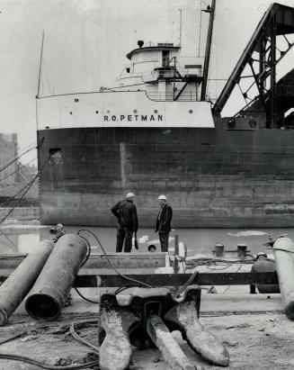 Image shows a big anchor lying by the lake with a ship in the background.