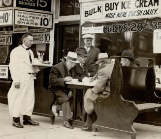 A waiter dressed in white jacket with black bow tie and long white apron holds a tray with tea …