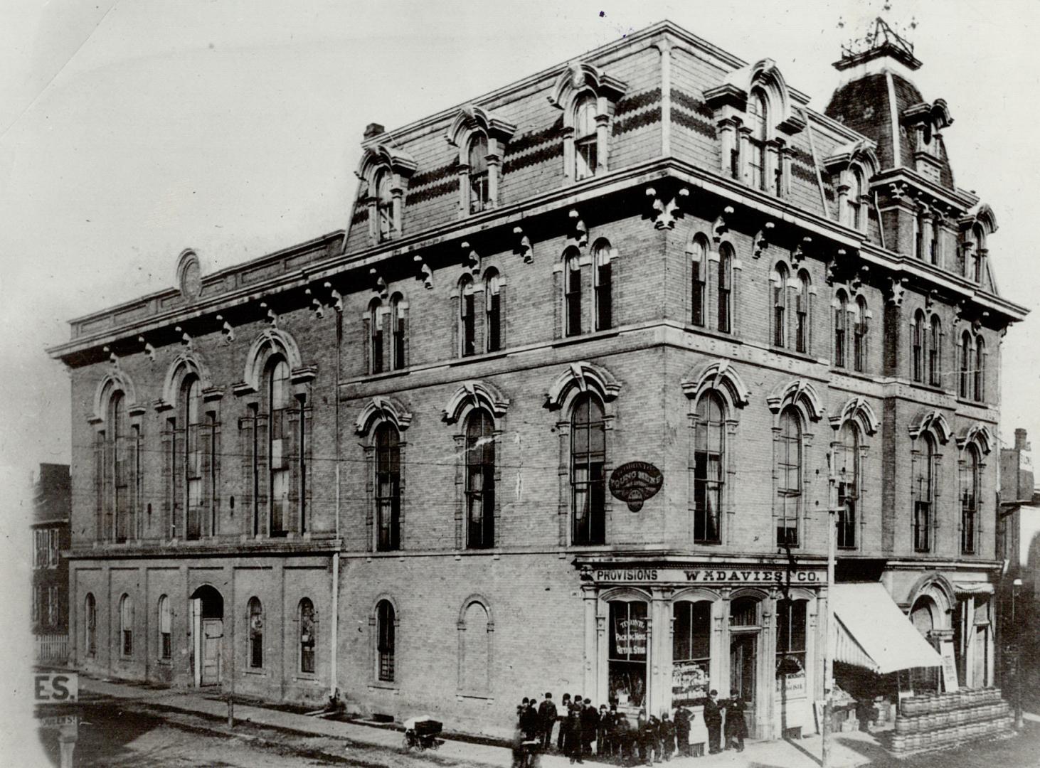 Corner view of four-storey brick building with Mansard roof. A group of men and boys dressed in…