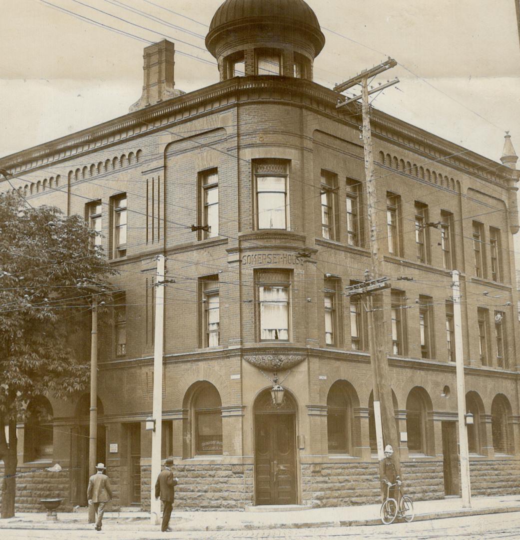 Corner view of three-storey brick building with turret. Policeman stands with bike at curb, rig…