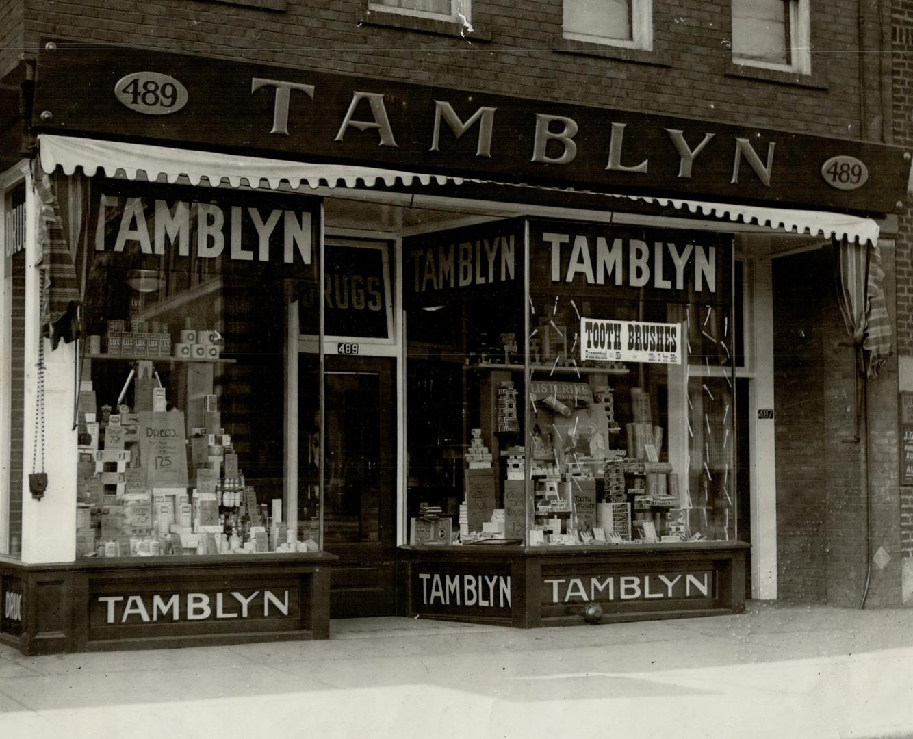 Storefront showing display windows full of merchandise either side of entrance. Sign above wind…