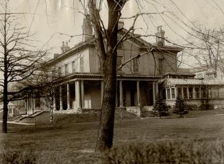 Two-storey structure with sloped roof, multiple brick chimneys, wraparound porch and attached s…