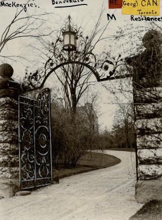 Chiseled stone posts support a decorative wrought iron arched gate at the entrance of a gravel …