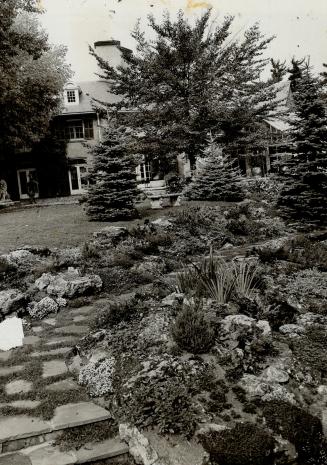 In foreground a flagstone path flanked by rock garden beds curves right, disappearing. Beyond, …