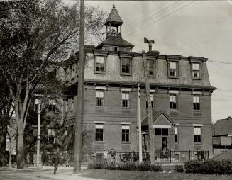 Three-storey brick building with belfry on roof and black fence surrounding. Children congregat…