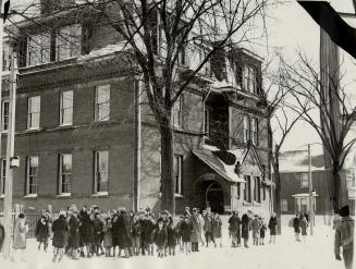 Three-storey brick building with Mansard roof and gabled portico entrance. Group of children an…