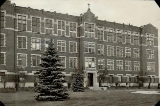 Four-storey brick building with flat roof. Cross stands on brick detail above roofline, centere…