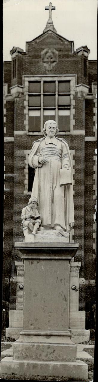 Carved, white stone statue of a man in gown and robe, holding paper in hand; left, a statue of …