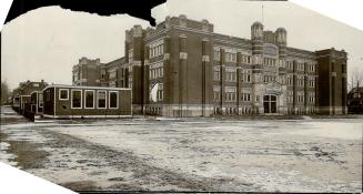 Exterior view of three-storey, flat roofed brick building looking onto flat, empty grounds. Sev…