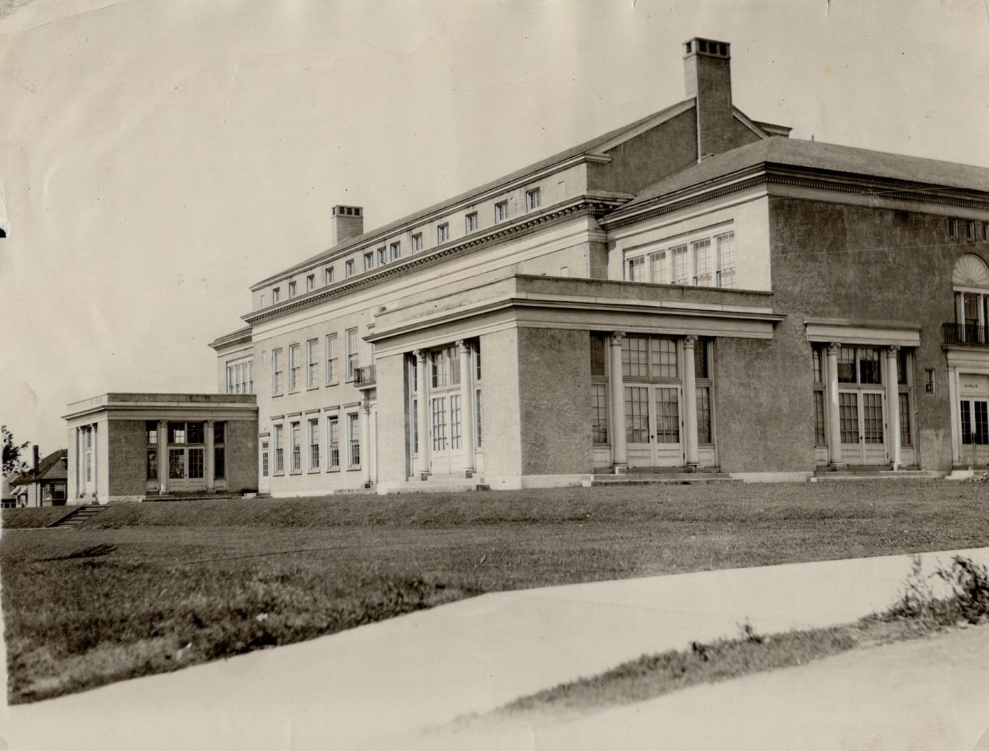 Exterior view of three-storey brick building on grassy incline, with tiered gabled wings of dou…