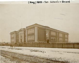 Exterior of three-storey brick building with flat roof, surrounded by an unpainted picket fence…