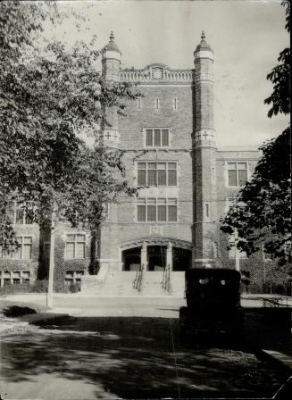 Narrow view of three-storey brick building with fourth floor tower above entrance. Right, in sh…