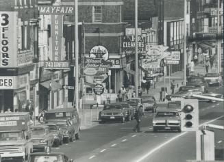 Street view of cars, trucks and vans parked next to sidewalk with business signs hanging overhe…