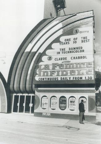 Man stands in front of five movie advertisements beside cinema entrance. Billboard above reads,…