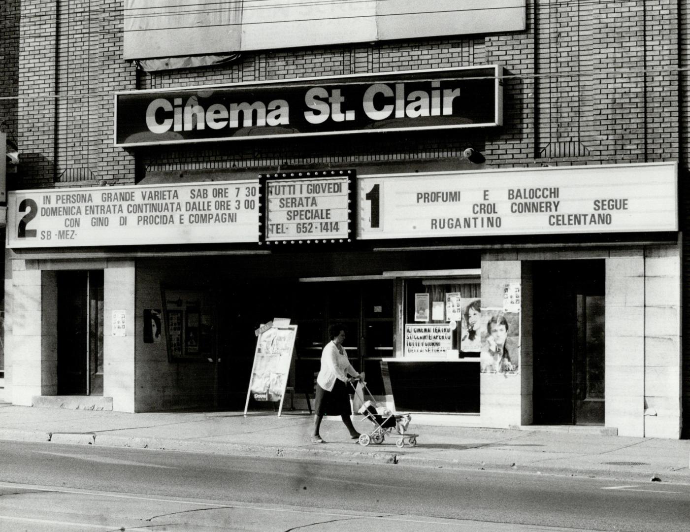 Woman passes with stroller in front of cinema entrance with glass ticket wiindow. Sign affixed …