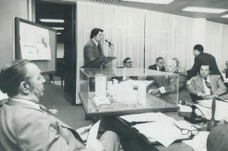 Image shows a person speaking into the microphone and others listening at the desks covered wit…